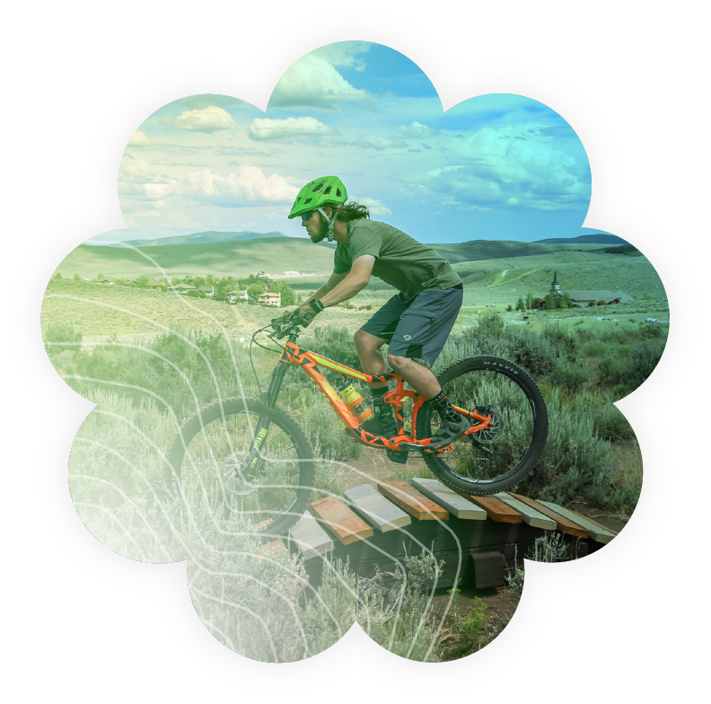 Decorative cover graphic featuring a mountain biker riding across a wooden bridge on a trail in Park City, Utah. The image is framed within a flower-shaped graphic overlay with topographic contour lines, representing outdoor recreation, trail systems, and adventure opportunities available through Basin Recreation.